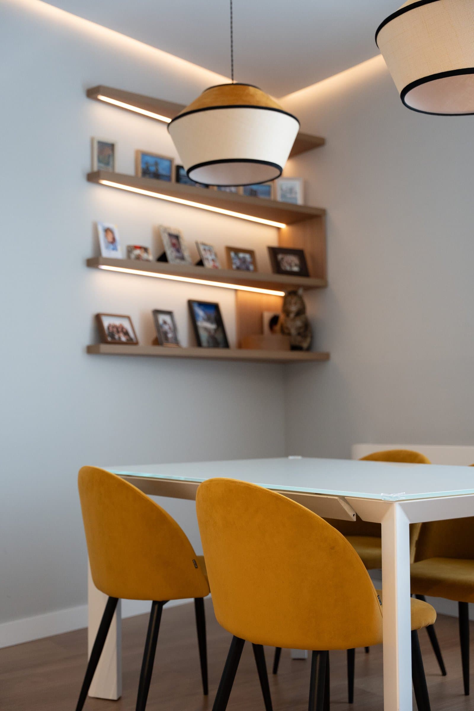 Dining table with mustard chairs under drum pendant lamps and lit picture shelves